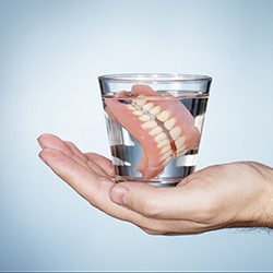 Hand holding full dentures in a glass of water