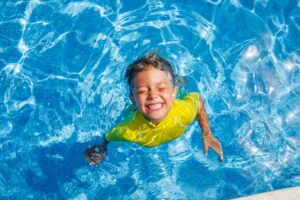 birds eye view of a young boy in a yellow shirt in a swimming pool looking up and smiling with eyes closed